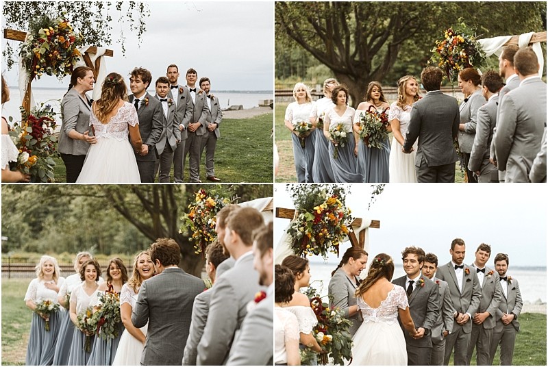 Premier Snohomish & Seattle Wedding Photographer | GSquared Weddings Photography A bride and groom stand together at a Snohomish outdoor wedding ceremony by the water, surrounded by bridesmaids in blue dresses and groomsmen in gray suits under a floral arch. Everyone looks joyful and engaged. 47.9129° N, 122.0982° W | Serving Snohomish, Seattle , Orlando & Beyond