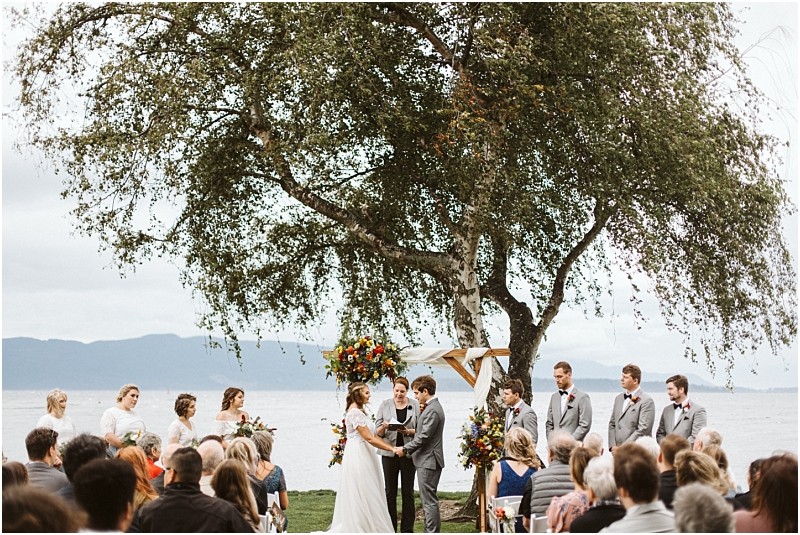 Premier Snohomish & Seattle Wedding Photographer | GSquared Weddings Photography A bride and groom stand under a floral arch by a large tree, exchanging vows at their Snohomish outdoor wedding ceremony by the water, surrounded by their wedding party and seated guests. 47.9129° N, 122.0982° W | Serving Snohomish, Seattle , Orlando & Beyond