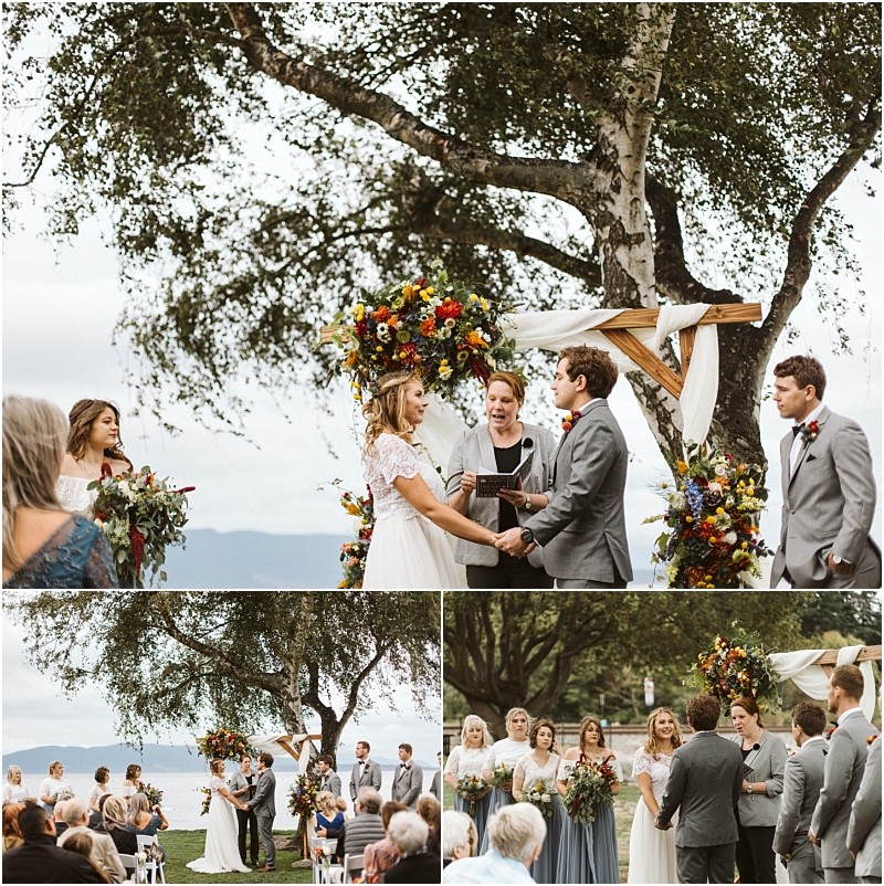 Premier Snohomish & Seattle Wedding Photographer | GSquared Weddings Photography A joyful couple exchanges vows under a floral arch outdoors at their Snohomish Seattle wedding, framed by a large tree. Guests are seated, bridesmaids in blue and groomsmen in gray stand by. The sky is overcast, adding romance to the celebration. 47.9129° N, 122.0982° W | Serving Snohomish, Seattle , Orlando & Beyond