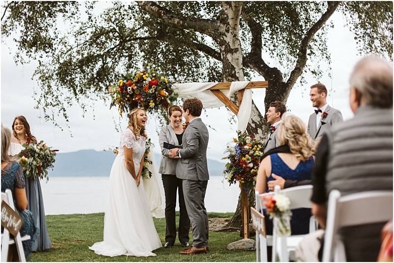 Premier Snohomish & Seattle Wedding Photographer | GSquared Weddings Photography A bride and groom stand smiling under a decorated wooden arch by a lake during an outdoor Snohomish wedding ceremony, surrounded by their wedding party and seated guests. 47.9129° N, 122.0982° W | Serving Snohomish, Seattle , Orlando & Beyond