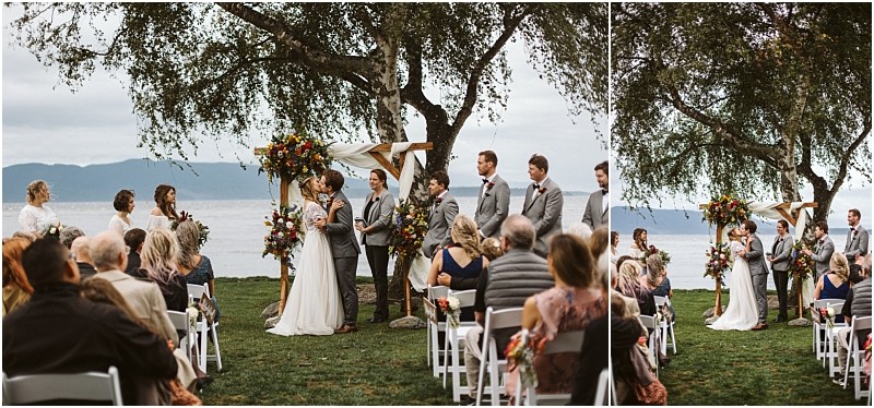 Premier Snohomish & Seattle Wedding Photographer | GSquared Weddings Photography A couple stands under a floral arch exchanging vows at an outdoor Snohomish lakeside wedding ceremony near Seattle, surrounded by their wedding party and guests on white chairs, with trees and water in the background. 47.9129° N, 122.0982° W | Serving Snohomish, Seattle , Orlando & Beyond