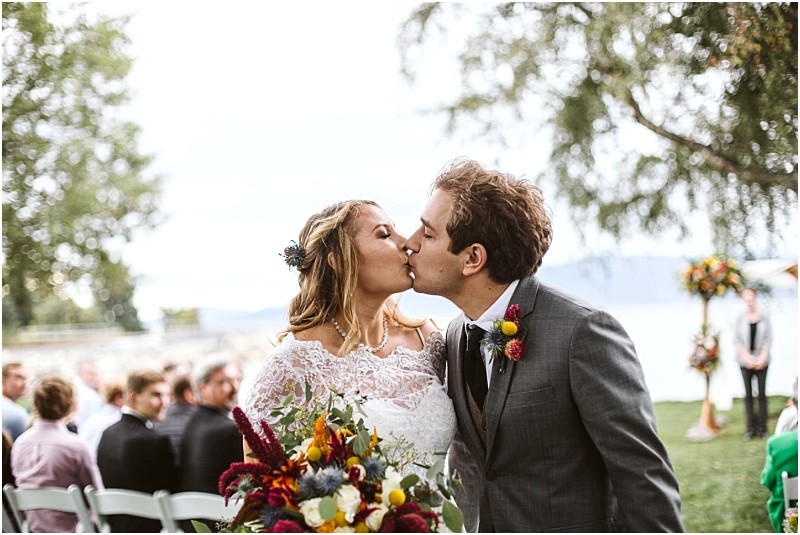 Premier Snohomish & Seattle Wedding Photographer | GSquared Weddings Photography A newlywed couple shares a kiss outdoors after their Snohomish wedding ceremony, holding a vibrant bouquet, with guests seated behind them and a lake and floral arch in the background. 47.9129° N, 122.0982° W | Serving Snohomish, Seattle , Orlando & Beyond
