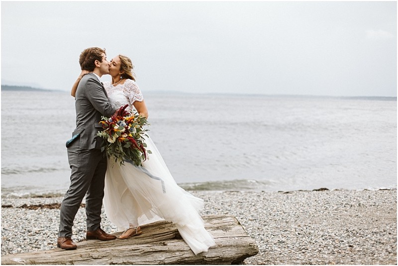 Premier Snohomish & Seattle Wedding Photographer | GSquared Weddings Photography A bride and groom kiss on a rocky beach near Snohomish, standing on driftwood. The bride holds a colorful bouquet in her white dress, while the groom wears a gray suit. The sea and cloudy sky set a romantic Seattle wedding scene. 47.9129° N, 122.0982° W | Serving Snohomish, Seattle , Orlando & Beyond