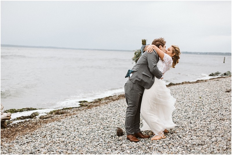 Premier Snohomish & Seattle Wedding Photographer | GSquared Weddings Photography A newlywed couple embraces and shares a kiss on a rocky Snohomish beach, with the ocean and cloudy sky in the background. The bride wears a flowing white dress and the groom is in a gray suit, capturing their romantic Seattle wedding. 47.9129° N, 122.0982° W | Serving Snohomish, Seattle , Orlando & Beyond