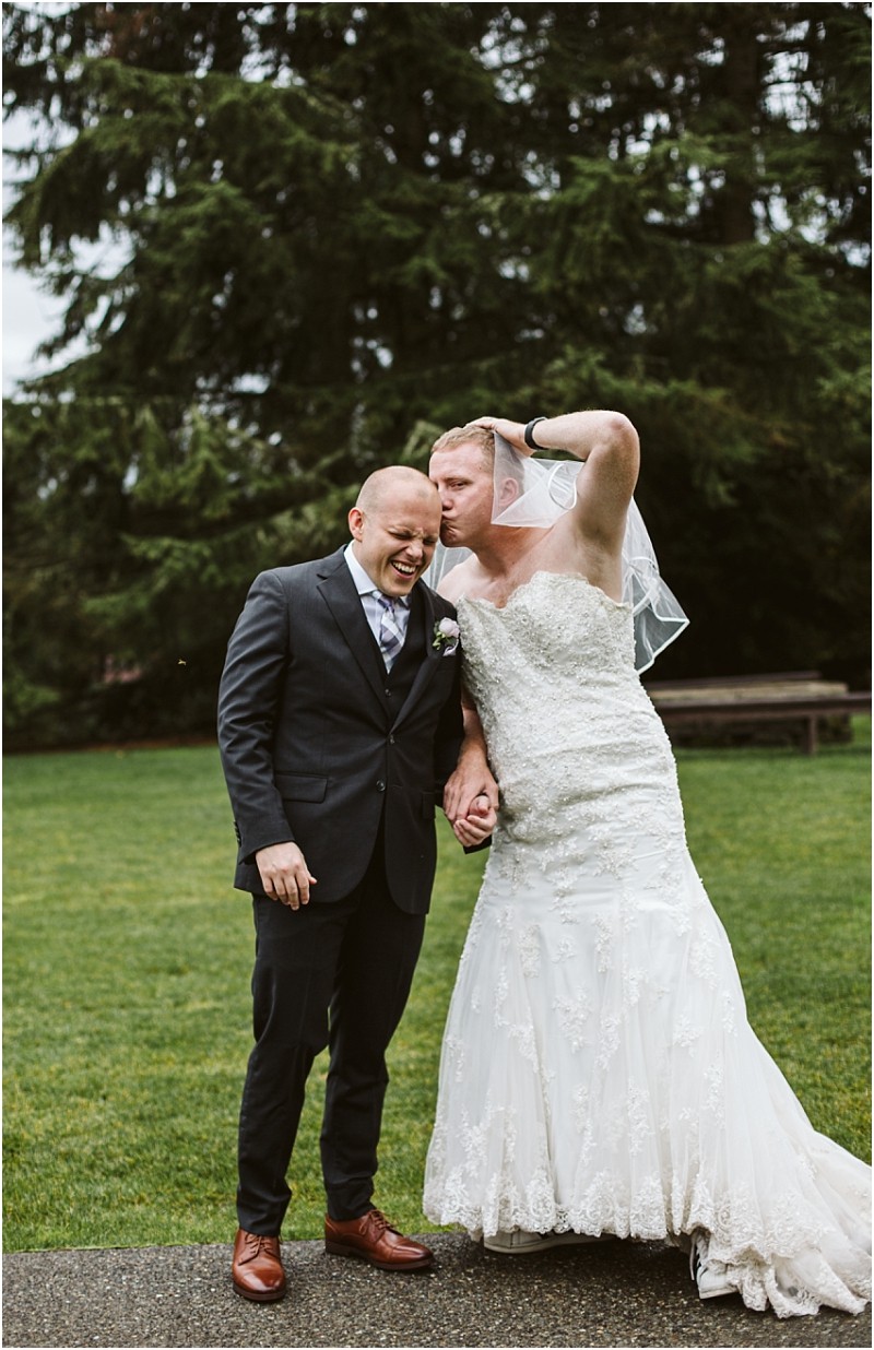 Premier Snohomish & Seattle Wedding Photographer | GSquared Weddings Photography Two people stand outside on grass at a Snohomish Seattle wedding. One, in a dark suit, laughs while holding hands with the other, who wears a white wedding dress and playfully flexes an arm muscle. Trees are visible in the background. 47.9129° N, 122.0982° W | Serving Snohomish, Seattle , Orlando & Beyond