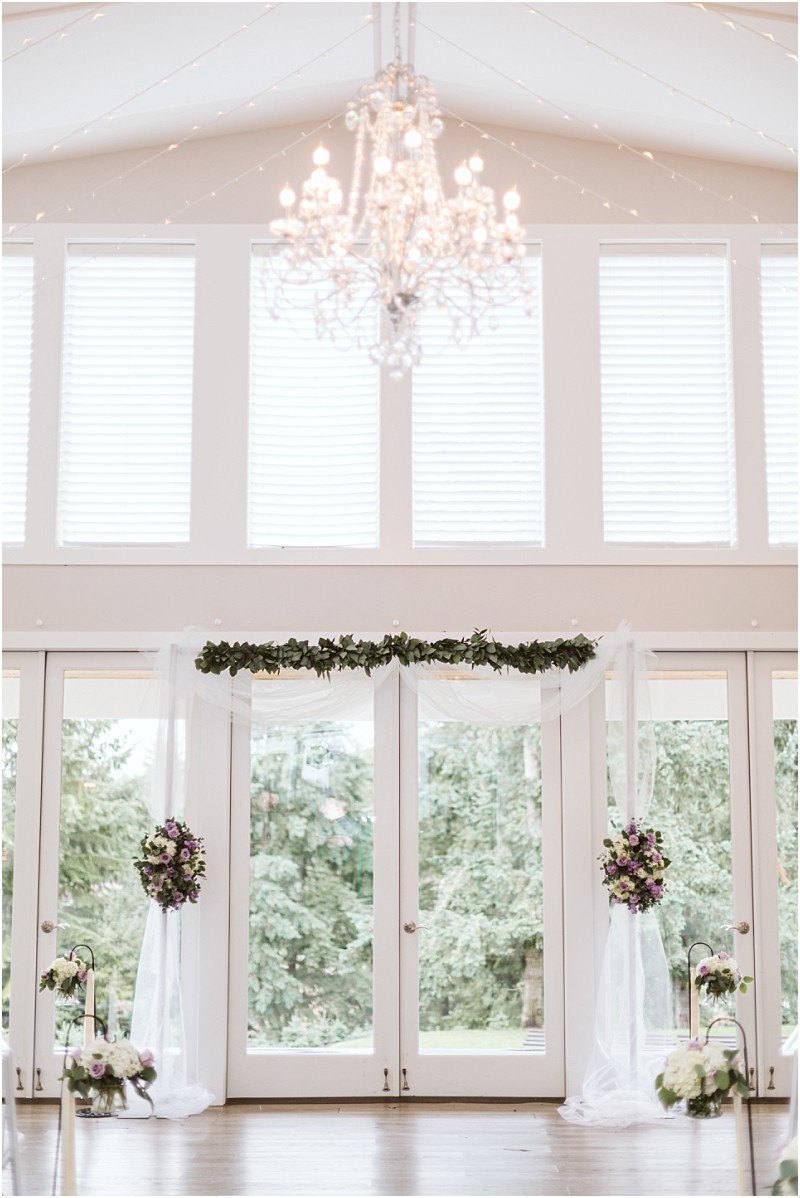 Premier Snohomish & Seattle Wedding Photographer | GSquared Weddings Photography Elegant indoor Snohomish wedding ceremony setup with tall windows, a sparkling chandelier, white drapery, green garland, and purple floral arrangements framing the glass doors, with trees visible outside. 47.9129° N, 122.0982° W | Serving Snohomish, Seattle , Orlando & Beyond