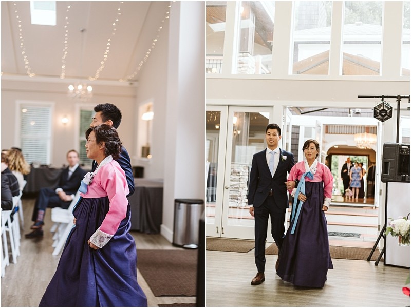Premier Snohomish & Seattle Wedding Photographer | GSquared Weddings Photography At a Snohomish Seattle wedding, a woman in a pink and navy hanbok walks beside a man in a suit at an indoor event, possibly an engagement, with guests seated and string lights glowing above. 47.9129° N, 122.0982° W | Serving Snohomish, Seattle , Orlando & Beyond