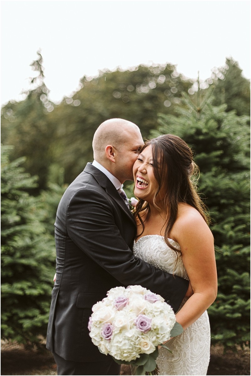Premier Snohomish & Seattle Wedding Photographer | GSquared Weddings Photography A groom in a black suit kisses his smiling bride, holding a bouquet of white and purple flowers. They stand close together outdoors, surrounded by green pine trees at their Snohomish Seattle wedding. 47.9129° N, 122.0982° W | Serving Snohomish, Seattle , Orlando & Beyond