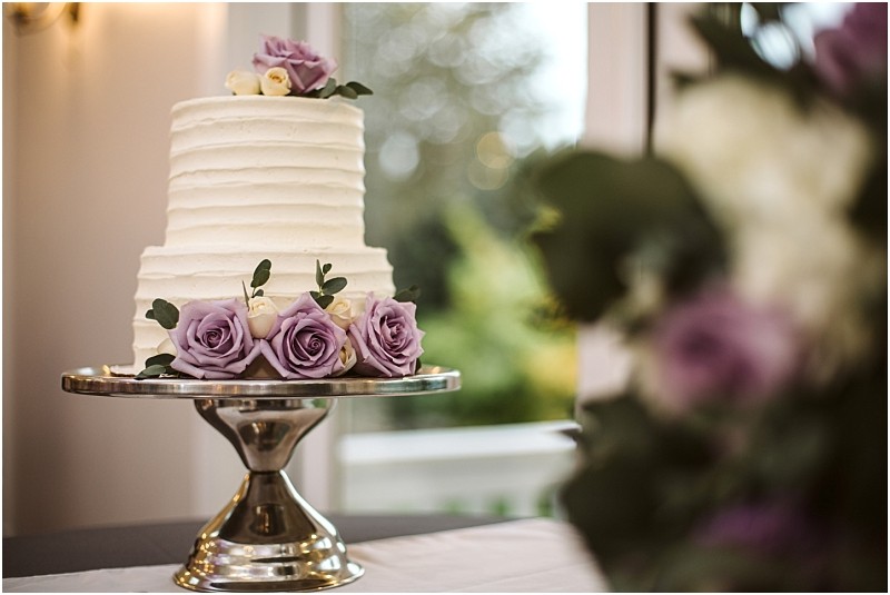 Premier Snohomish & Seattle Wedding Photographer | GSquared Weddings Photography A two-tiered white cake decorated with purple and white roses sits on a silver cake stand—a perfect centerpiece for a Snohomish or Seattle wedding. The softly blurred background reveals lush greenery through a window. 47.9129° N, 122.0982° W | Serving Snohomish, Seattle , Orlando & Beyond