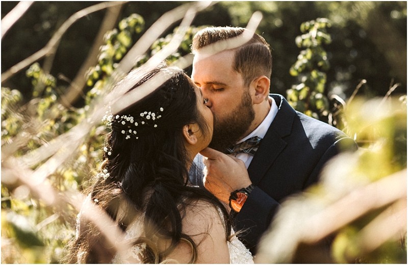 Premier Snohomish & Seattle Wedding Photographer | GSquared Weddings Photography A bride and groom share a kiss outdoors at their Snohomish Seattle wedding, surrounded by greenery. The bride’s dark hair is adorned with small white flowers, and the groom in a navy suit gently holds her chin as sunlight filters through branches. 47.9129° N, 122.0982° W | Serving Snohomish, Seattle , Orlando & Beyond
