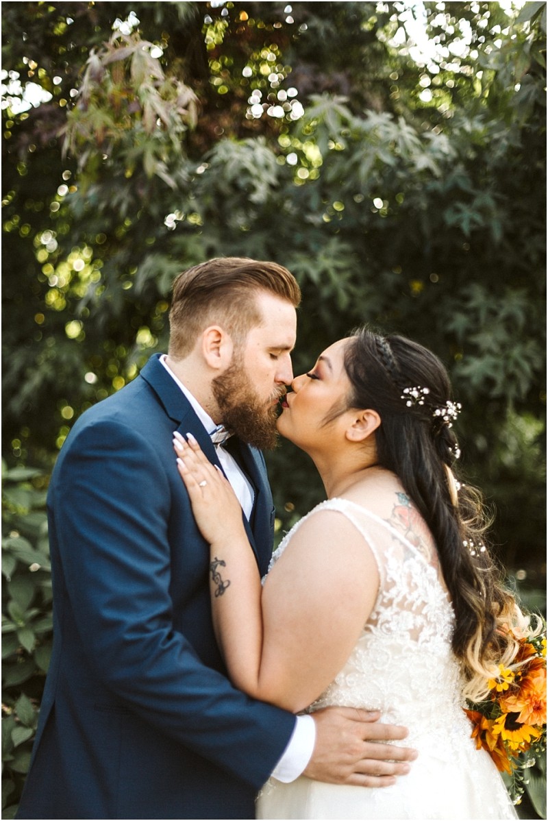 Premier Snohomish & Seattle Wedding Photographer | GSquared Weddings Photography A bride and groom, dressed in a white lace wedding dress and navy suit, lovingly kiss outdoors in front of lush green trees. Capturing a magical Snohomish Seattle wedding moment, the bride holds a bouquet and has flowers in her hair. 47.9129° N, 122.0982° W | Serving Snohomish, Seattle , Orlando & Beyond