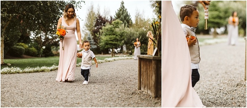 Premier Snohomish & Seattle Wedding Photographer | GSquared Weddings Photography A woman in a pink dress holding orange flowers walks outdoors on a gravel path with a young boy in a vest and bowtie, slightly ahead of her. Lush greenery and other guests set the scene for this charming Snohomish Seattle wedding moment. 47.9129° N, 122.0982° W | Serving Snohomish, Seattle , Orlando & Beyond