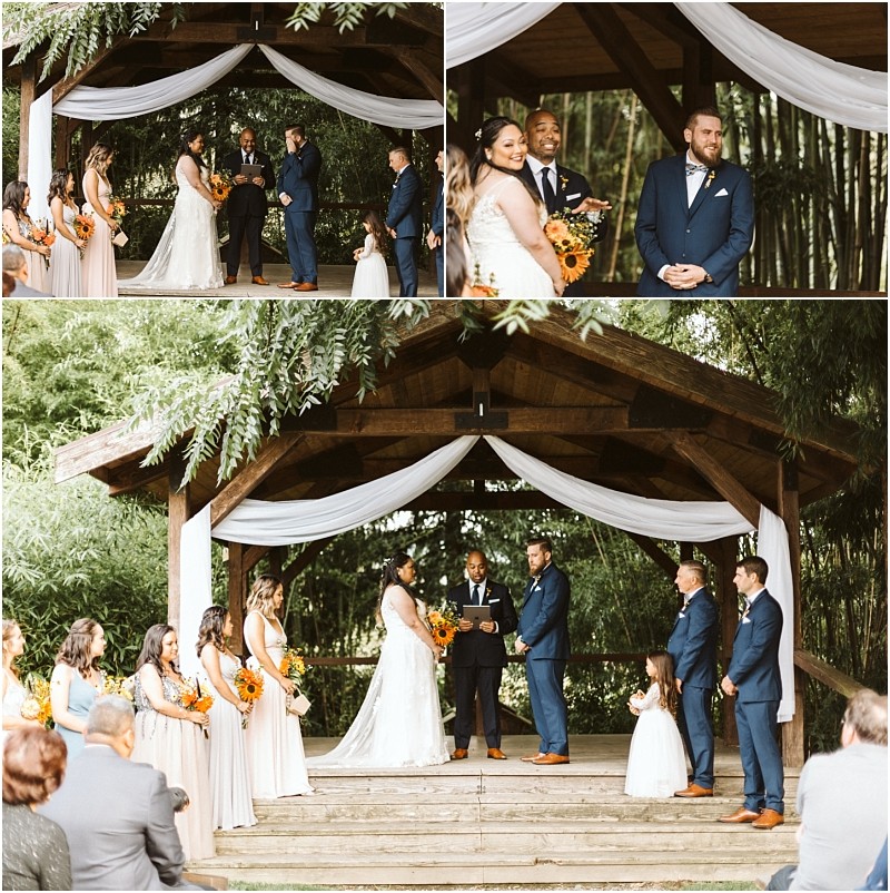 Premier Snohomish & Seattle Wedding Photographer | GSquared Weddings Photography A romantic Snohomish wedding ceremony unfolds outdoors under a wooden pavilion with white drapery. The bride and groom stand facing each other, surrounded by their wedding party and guests, with lush greenery enhancing this Seattle engagement moment. 47.9129° N, 122.0982° W | Serving Snohomish, Seattle , Orlando & Beyond