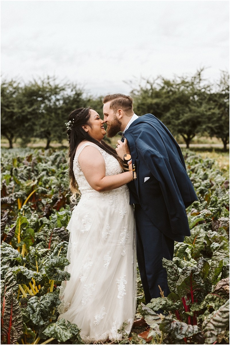 Premier Snohomish & Seattle Wedding Photographer | GSquared Weddings Photography A bride and groom stand closely together in a lush Snohomish vegetable garden, smiling and about to kiss. The bride wears a white lace gown, and the groom, in a navy suit, drapes his jacket over his shoulder. Trees frame this Seattle wedding moment. 47.9129° N, 122.0982° W | Serving Snohomish, Seattle , Orlando & Beyond