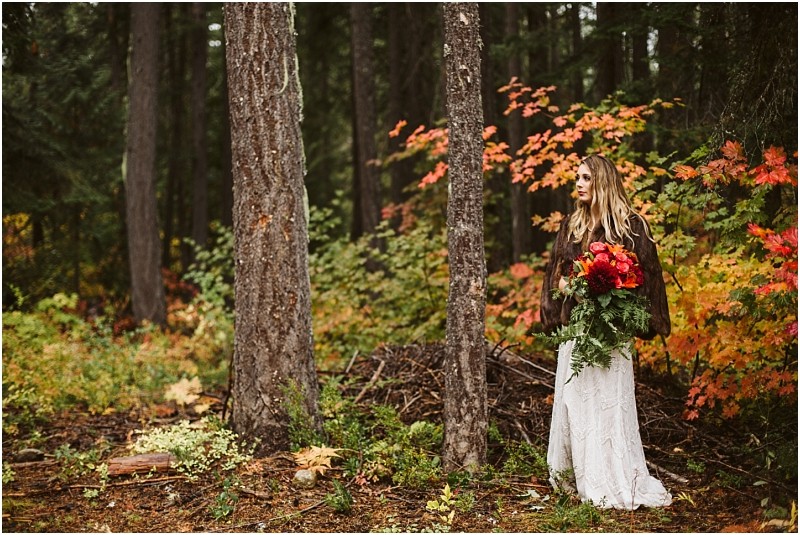 Premier Snohomish & Seattle Wedding Photographer | GSquared Weddings Photography A woman in a white dress and brown jacket stands in a forest near Snohomish, holding a bouquet of red and orange flowers. Vibrant autumn leaves surround her, with tall trees in the background—perfect for a Seattle wedding engagement shoot. 47.9129° N, 122.0982° W | Serving Snohomish, Seattle , Orlando & Beyond