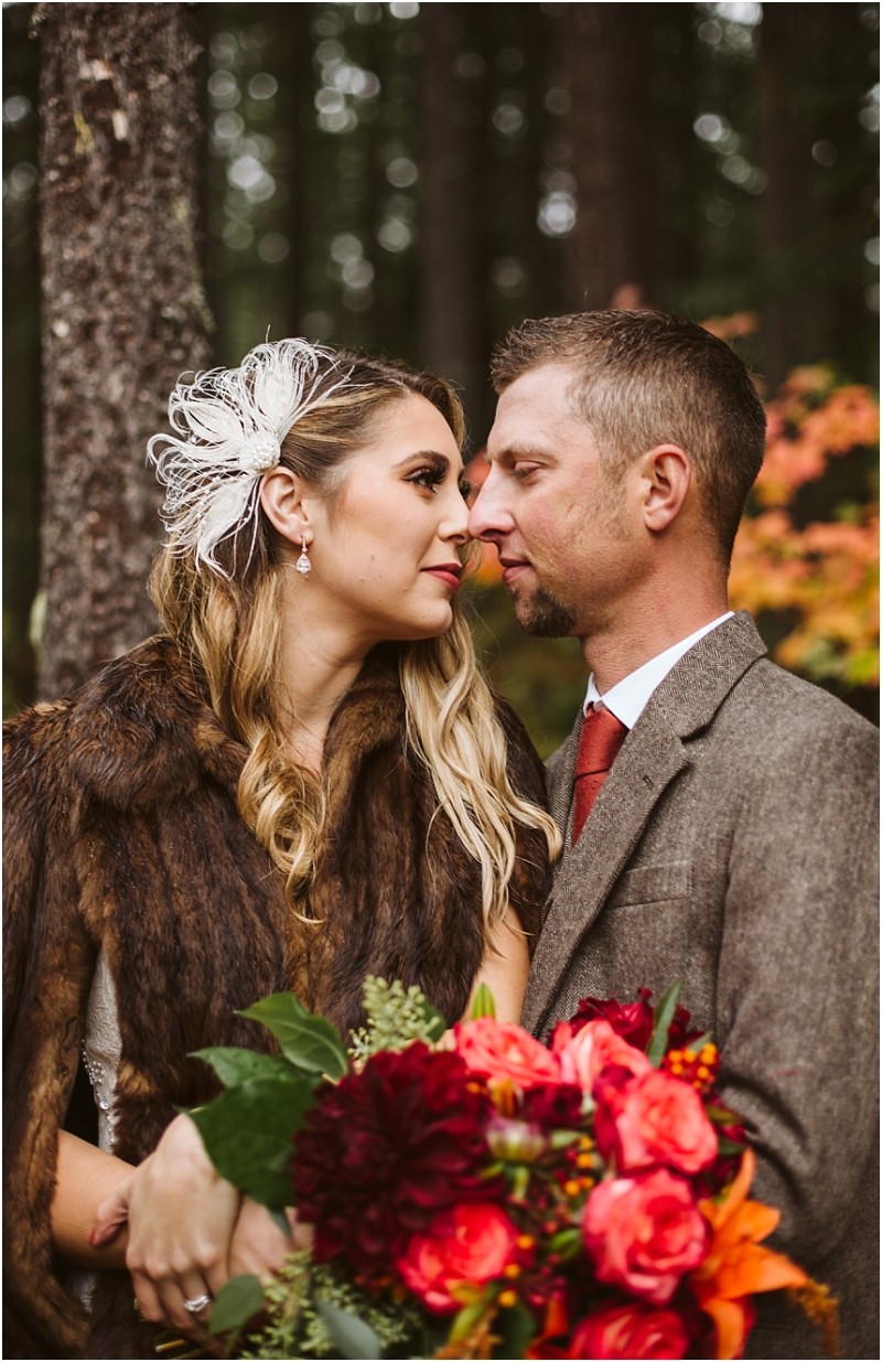 Premier Snohomish & Seattle Wedding Photographer | GSquared Weddings Photography A bride and groom stand close together in a forest during their Snohomish wedding; the bride wears a fur shawl, feather hair accessory, and holds a vibrant bouquet. The groom complements her with a brown suit and red tie. 47.9129° N, 122.0982° W | Serving Snohomish, Seattle , Orlando & Beyond