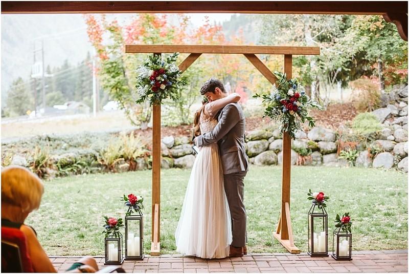 Premier Snohomish & Seattle Wedding Photographer | GSquared Weddings Photography A bride and groom embrace under a wooden wedding arch decorated with flowers and lanterns outdoors, with greenery and autumn foliage in the background at a Snohomish Seattle wedding. A seated guest is partially visible in the foreground. 47.9129° N, 122.0982° W | Serving Snohomish, Seattle , Orlando & Beyond
