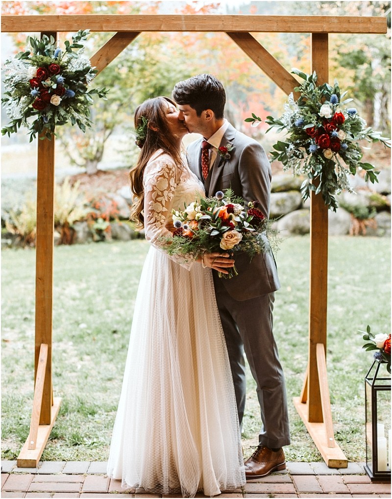 Premier Snohomish & Seattle Wedding Photographer | GSquared Weddings Photography A bride and groom kiss under a wooden arch decorated with flowers in a picturesque Snohomish outdoor setting. The bride holds a bouquet in her lace dress, while the groom wears a gray suit and patterned tie. 47.9129° N, 122.0982° W | Serving Snohomish, Seattle , Orlando & Beyond