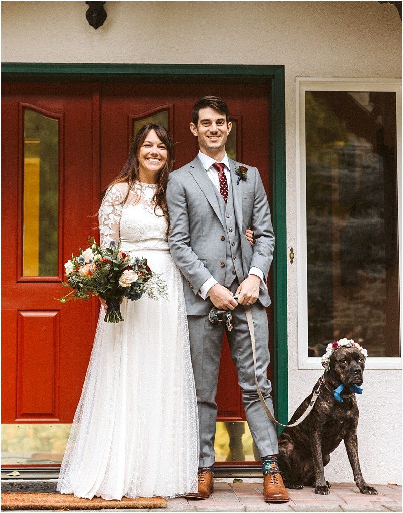 Premier Snohomish & Seattle Wedding Photographer | GSquared Weddings Photography A smiling bride in a white lace dress stands beside a groom in a gray suit holding a leash. Their dog, wearing a flower crown, sits near them in front of a red door at their dreamy Snohomish Seattle wedding. 47.9129° N, 122.0982° W | Serving Snohomish, Seattle , Orlando & Beyond