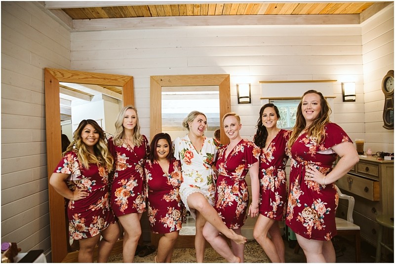 Premier Snohomish & Seattle Wedding Photographer | GSquared Weddings Photography Seven women pose together in a room with wooden walls and mirrors during a Snohomish Seattle wedding; six wear matching red floral robes, while the bride in white playfully lifts her leg as everyone smiles at the camera. 47.9129° N, 122.0982° W | Serving Snohomish, Seattle , Orlando & Beyond