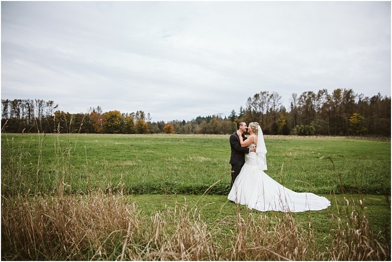 Premier Snohomish & Seattle Wedding Photographer | GSquared Weddings Photography A bride and groom stand together in an embrace on a grassy field near Snohomish, surrounded by tall grass and trees under an overcast sky. The bride wears a white gown and veil, while the groom is in a dark suit—capturing their Seattle wedding magic. 47.9129° N, 122.0982° W | Serving Snohomish, Seattle , Orlando & Beyond