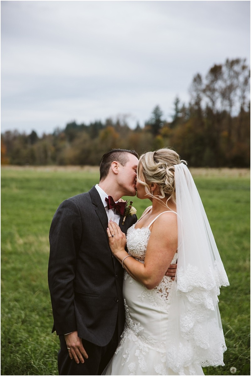 Premier Snohomish & Seattle Wedding Photographer | GSquared Weddings Photography A bride and groom share a kiss outdoors in a green field near Snohomish, with trees and a cloudy sky in the background. The bride wears a white dress and veil, while the groom is in a dark suit with a bow tie. 47.9129° N, 122.0982° W | Serving Snohomish, Seattle , Orlando & Beyond
