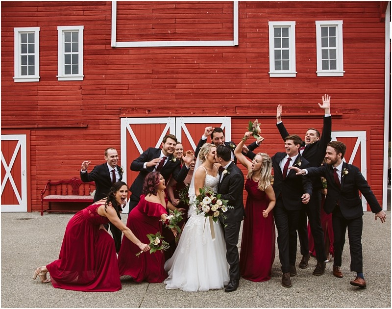 Premier Snohomish & Seattle Wedding Photographer | GSquared Weddings Photography A joyful Snohomish wedding party poses in front of a red barn. The bride and groom share a kiss, surrounded by bridesmaids in red dresses and groomsmen in dark suits. Everyone is smiling and celebrating this special Seattle engagement. 47.9129° N, 122.0982° W | Serving Snohomish, Seattle , Orlando & Beyond