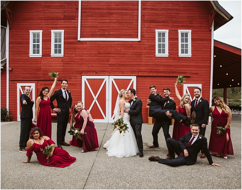 Premier Snohomish & Seattle Wedding Photographer | GSquared Weddings Photography A bride and groom share a kiss in front of a red barn at their Snohomish wedding, surrounded by their joyful party in burgundy attire, posing playfully and holding bouquets. 47.9129° N, 122.0982° W | Serving Snohomish, Seattle , Orlando & Beyond