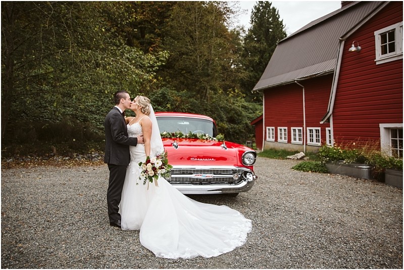 Premier Snohomish & Seattle Wedding Photographer | GSquared Weddings Photography A bride and groom share a kiss in front of a classic red Chevrolet at their Snohomish wedding, parked beside a red barn and surrounded by trees. The bride holds a bouquet and wears a white gown with a long train. 47.9129° N, 122.0982° W | Serving Snohomish, Seattle , Orlando & Beyond
