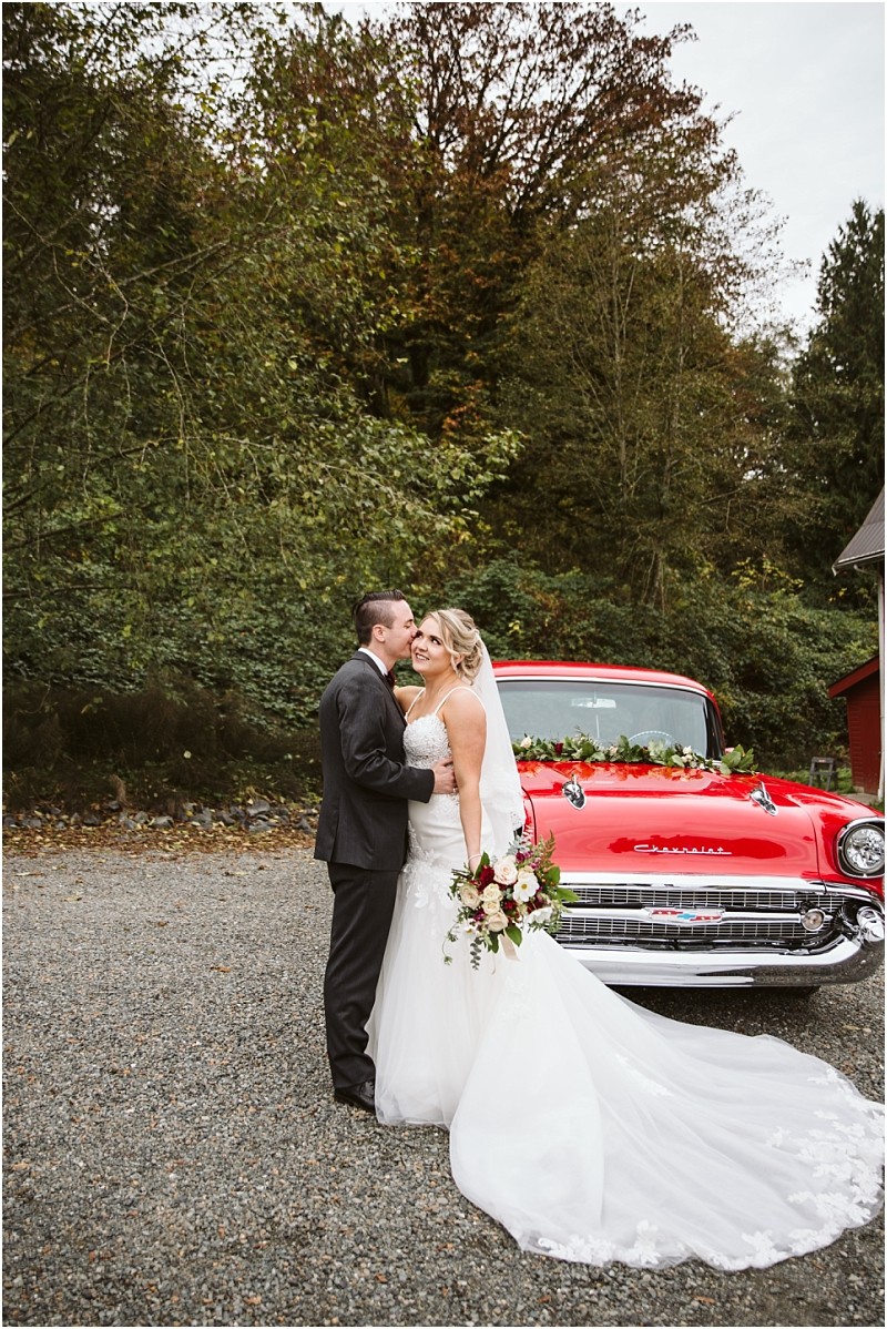 Premier Snohomish & Seattle Wedding Photographer | GSquared Weddings Photography A bride and groom embrace and smile in front of a vintage red car decorated with flowers at their Snohomish Seattle wedding, surrounded by trees and greenery on a gravel driveway. 47.9129° N, 122.0982° W | Serving Snohomish, Seattle , Orlando & Beyond