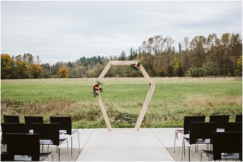 Premier Snohomish & Seattle Wedding Photographer | GSquared Weddings Photography A wooden hexagonal wedding arch decorated with flowers stands outdoors on a patio in Snohomish, facing a grassy field and trees. Black chairs are arranged for guests under a cloudy Seattle sky, perfect for a wedding or engagement ceremony. 47.9129° N, 122.0982° W | Serving Snohomish, Seattle , Orlando & Beyond