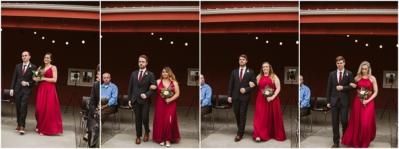 Premier Snohomish & Seattle Wedding Photographer | GSquared Weddings Photography Four pairs of bridesmaids in red dresses and groomsmen in dark suits walk arm-in-arm outdoors at a Snohomish Seattle wedding, with a red barn and seated guests in the background. 47.9129° N, 122.0982° W | Serving Snohomish, Seattle , Orlando & Beyond