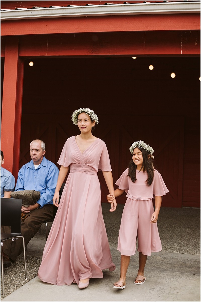 Premier Snohomish & Seattle Wedding Photographer | GSquared Weddings Photography Two girls in matching dusty pink dresses and flower crowns walk hand in hand outdoors, smiling, at a Snohomish wedding. A man in blue sits on a chair to their left, watching them pass by the red building. 47.9129° N, 122.0982° W | Serving Snohomish, Seattle , Orlando & Beyond