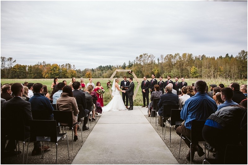 Premier Snohomish & Seattle Wedding Photographer | GSquared Weddings Photography An outdoor Snohomish wedding ceremony features a bride and groom standing under an arch, surrounded by their wedding party. Guests are seated on both sides, with trees and fields in the background under a cloudy sky. 47.9129° N, 122.0982° W | Serving Snohomish, Seattle , Orlando & Beyond