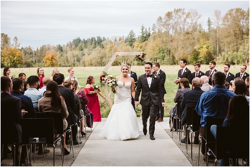 Premier Snohomish & Seattle Wedding Photographer | GSquared Weddings Photography A bride and groom walk down the aisle outside at their Snohomish wedding, smiling and holding hands, surrounded by seated guests and a joyful wedding party. Lush greenery and towering trees create the perfect Seattle engagement backdrop. 47.9129° N, 122.0982° W | Serving Snohomish, Seattle , Orlando & Beyond