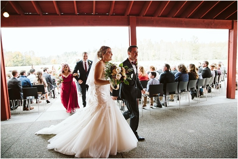 Premier Snohomish & Seattle Wedding Photographer | GSquared Weddings Photography A bride and groom walk hand in hand, smiling, after their Snohomish wedding ceremony, followed by the bridal party. Guests seated outdoors watch under a covered patio with a scenic background near Seattle. 47.9129° N, 122.0982° W | Serving Snohomish, Seattle , Orlando & Beyond
