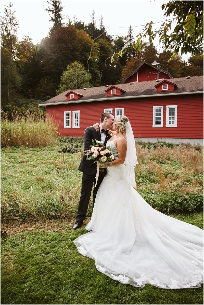 Premier Snohomish & Seattle Wedding Photographer | GSquared Weddings Photography A bride and groom share a kiss outdoors in front of a bright red barn near Snohomish, surrounded by greenery. The bride wears a white gown with a long train and veil, while the groom is in a dark suit at their Seattle wedding. 47.9129° N, 122.0982° W | Serving Snohomish, Seattle , Orlando & Beyond