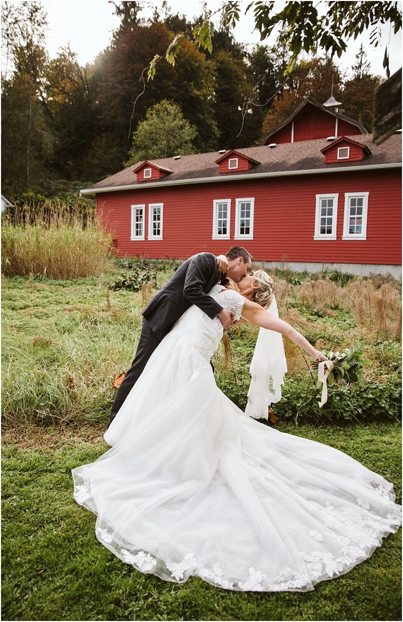 Premier Snohomish & Seattle Wedding Photographer | GSquared Weddings Photography A groom in a black suit dips and kisses his bride in a white wedding gown outdoors by a red building, with greenery and trees in the background—a romantic Snohomish Seattle wedding moment. 47.9129° N, 122.0982° W | Serving Snohomish, Seattle , Orlando & Beyond