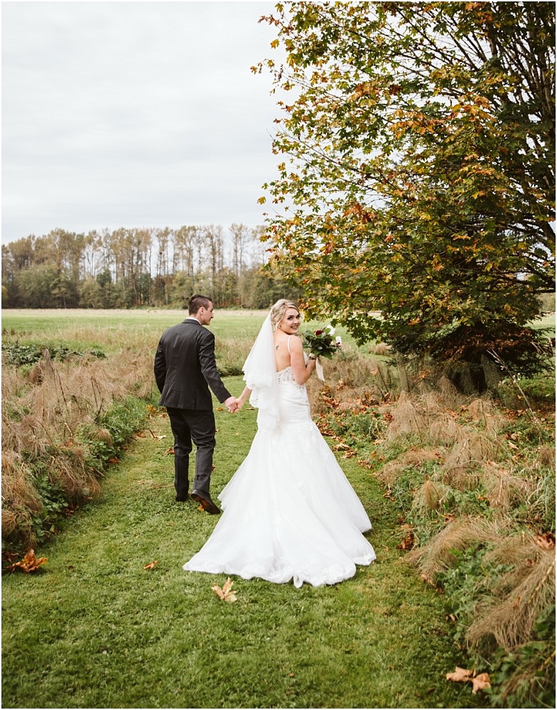 Premier Snohomish & Seattle Wedding Photographer | GSquared Weddings Photography A bride in a white gown and veil holds a bouquet and looks back, smiling, while walking hand-in-hand with her groom in a dark suit along a grassy path surrounded by autumn trees—capturing a magical Snohomish Seattle wedding moment. 47.9129° N, 122.0982° W | Serving Snohomish, Seattle , Orlando & Beyond