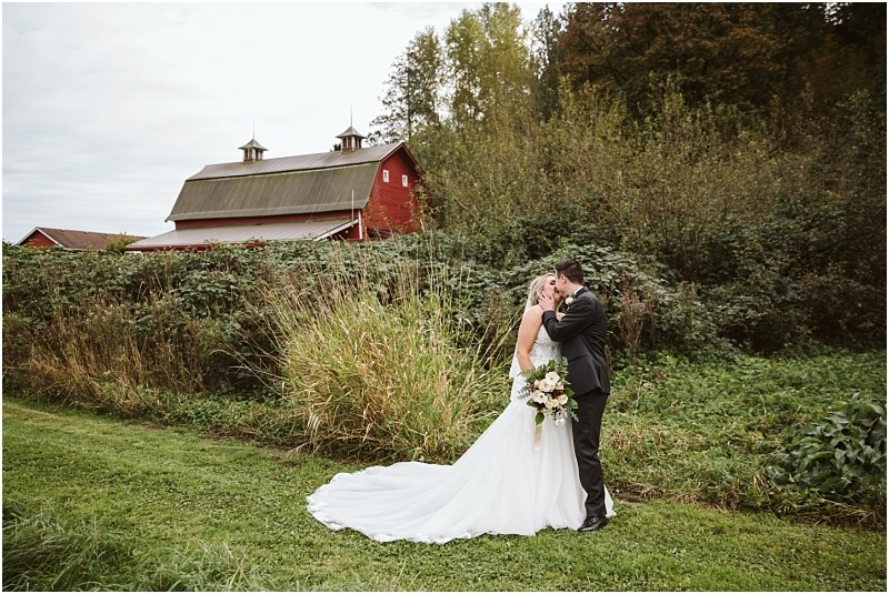 Premier Snohomish & Seattle Wedding Photographer | GSquared Weddings Photography A Snohomish bride and groom embrace and kiss outside on a grassy lawn, with a red barn and tall green bushes in the background. The bride wears a white gown and holds a bouquet; the groom is in a black suit. 47.9129° N, 122.0982° W | Serving Snohomish, Seattle , Orlando & Beyond