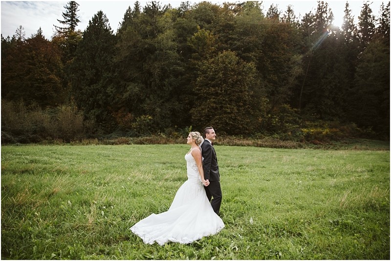 Premier Snohomish & Seattle Wedding Photographer | GSquared Weddings Photography A bride and groom stand back-to-back holding hands in a grassy field near Snohomish, surrounded by tall trees and greenery, with sunlight filtering through the forest in the background—a perfect Seattle wedding moment. 47.9129° N, 122.0982° W | Serving Snohomish, Seattle , Orlando & Beyond