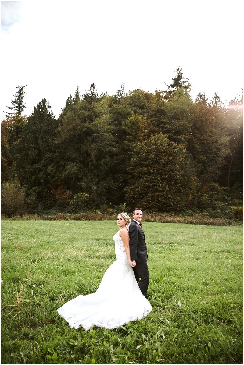 Premier Snohomish & Seattle Wedding Photographer | GSquared Weddings Photography A bride in a white gown and a groom in a dark suit stand back-to-back on a grassy field, framed by trees and a bright sky, capturing the essence of a romantic Snohomish Seattle wedding engagement. 47.9129° N, 122.0982° W | Serving Snohomish, Seattle , Orlando & Beyond