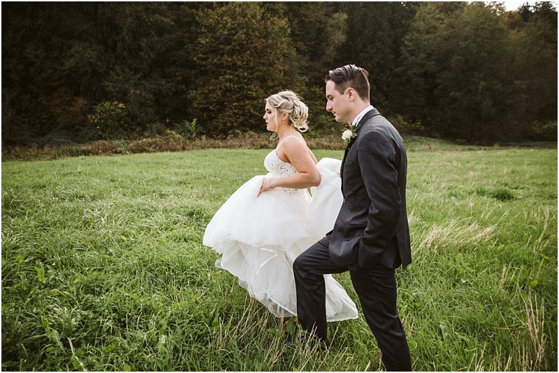Premier Snohomish & Seattle Wedding Photographer | GSquared Weddings Photography A bride in a white gown and a groom in a dark suit walk together through a grassy field with trees in the background, capturing the charm of a Snohomish Seattle wedding as the bride lifts her dress while they move forward. 47.9129° N, 122.0982° W | Serving Snohomish, Seattle , Orlando & Beyond
