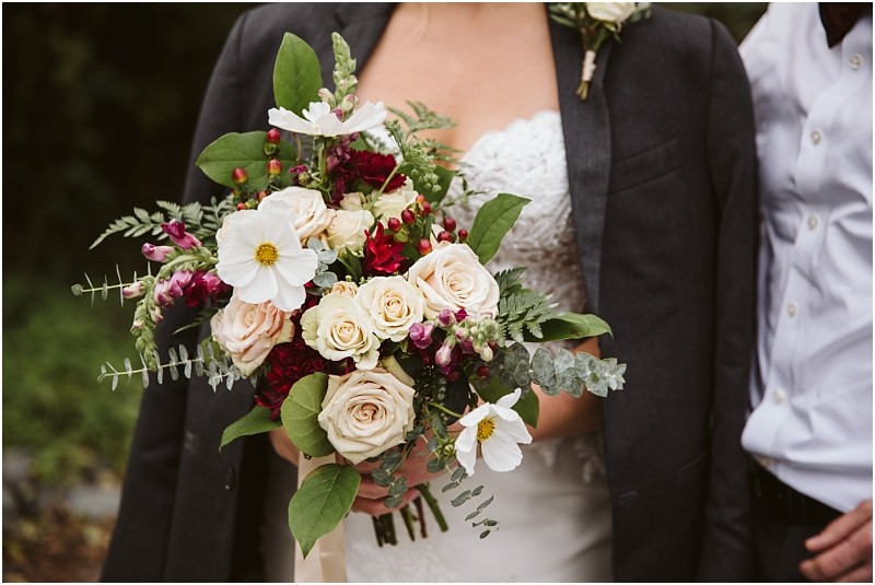 Premier Snohomish & Seattle Wedding Photographer | GSquared Weddings Photography A bride in a strapless white dress and dark blazer holds a bouquet of cream roses, white daisies, and greenery at a Snohomish Seattle wedding. Another person in a white shirt stands beside her, both partially visible. 47.9129° N, 122.0982° W | Serving Snohomish, Seattle , Orlando & Beyond