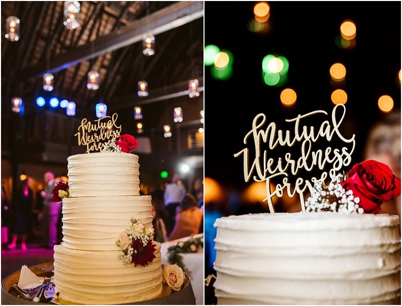 Premier Snohomish & Seattle Wedding Photographer | GSquared Weddings Photography A white tiered wedding cake decorated with red and white flowers, featuring a gold topper that reads “Mutual Weirdness Forever,” is displayed at a dimly lit Snohomish wedding reception with hanging lights. 47.9129° N, 122.0982° W | Serving Snohomish, Seattle , Orlando & Beyond