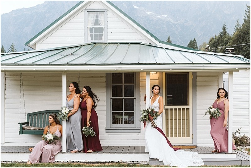 Premier Snohomish & Seattle Wedding Photographer | GSquared Weddings Photography Five women in formal dresses, including a bride in white holding a bouquet, pose on the porch of a white house with a green roof near Snohomish, set against a scenic mountain and tree backdrop. 47.9129° N, 122.0982° W | Serving Snohomish, Seattle , Orlando & Beyond