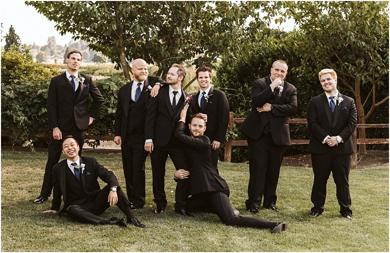 Premier Snohomish & Seattle Wedding Photographer | GSquared Weddings Photography A group of eight men in black suits and ties pose playfully outdoors on grass at a Snohomish Seattle wedding, with trees and a wooden fence in the background. Some stand smiling, while others strike humorous, relaxed poses in front. 47.9129° N, 122.0982° W | Serving Snohomish, Seattle , Orlando & Beyond