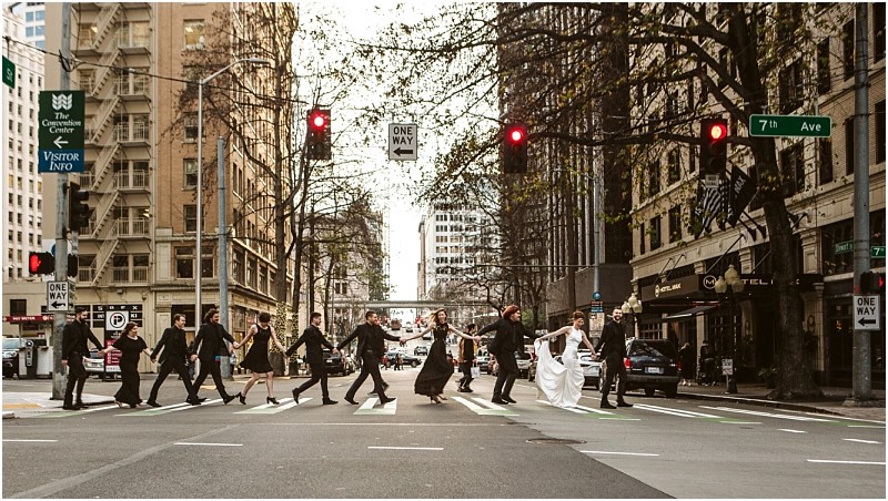 Premier Snohomish & Seattle Wedding Photographer | GSquared Weddings Photography A wedding party crosses a city street at a crosswalk, hand in hand, with the bride and groom at the center, surrounded by attendants in formal black attire. Tall buildings line the scene during this unforgettable Seattle wedding. 47.9129° N, 122.0982° W | Serving Snohomish, Seattle , Orlando & Beyond