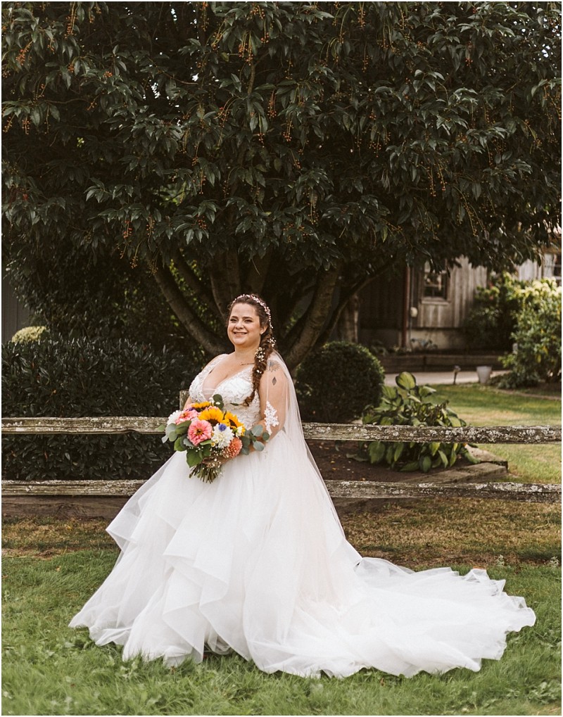 Premier Snohomish & Seattle Wedding Photographer | GSquared Weddings Photography A bride in a white, layered wedding gown stands on grass near Snohomish, smiling and holding a vibrant bouquet. She has long, braided hair and is posed in front of a large leafy tree with rustic buildings in the background. 47.9129° N, 122.0982° W | Serving Snohomish, Seattle , Orlando & Beyond