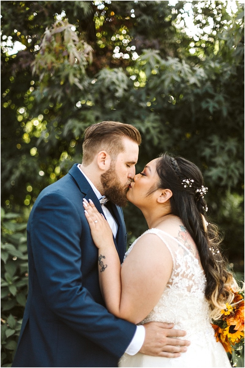 Premier Snohomish & Seattle Wedding Photographer | GSquared Weddings Photography A Snohomish bride and groom share a kiss outdoors, surrounded by greenery. The groom wears a navy suit and the bride has a lace dress with flowers in her hair, holding a bouquet. Sunlight filters through the leaves behind them. 47.9129° N, 122.0982° W | Serving Snohomish, Seattle , Orlando & Beyond